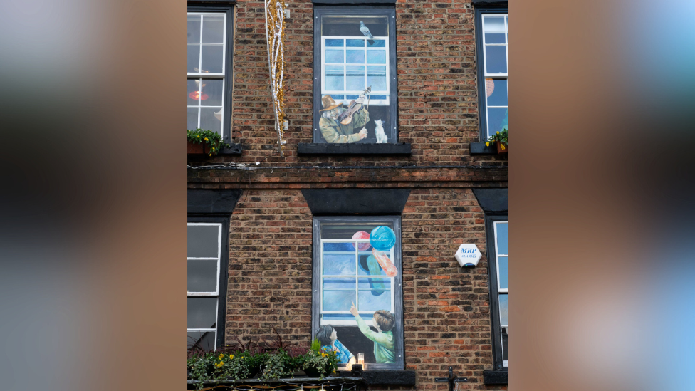 A top-floor window painting depicts a man with a cat and a fiddle. In a window painting directly below, two people point up towards the man.