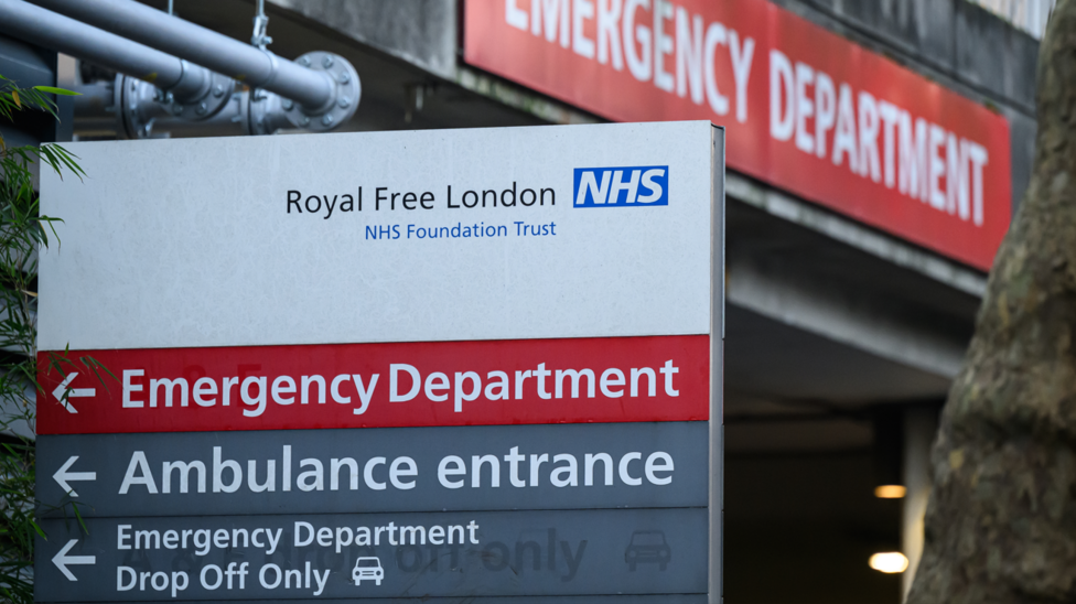 A white sign outside the hospital reads "Royal Free London", with a red strip underneath saying "Emergency Department" in white lettering, and grey strips below saying "Ambulance entrance" and "Emergency Department Drop Off Only" in white lettering, with a white symbol of a car. In the background a red sign with white lettering marks the Emergency Department entrance