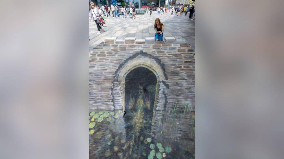 Artwork showing a water gate on the ground drawn in chalk. There is a water leading up to the gate with lily pads on the water in the art. A woman kneels down to admire the art at the top of the photo. The optical illusion makes it look like the girl is sat on top of the wall of the water gate. 