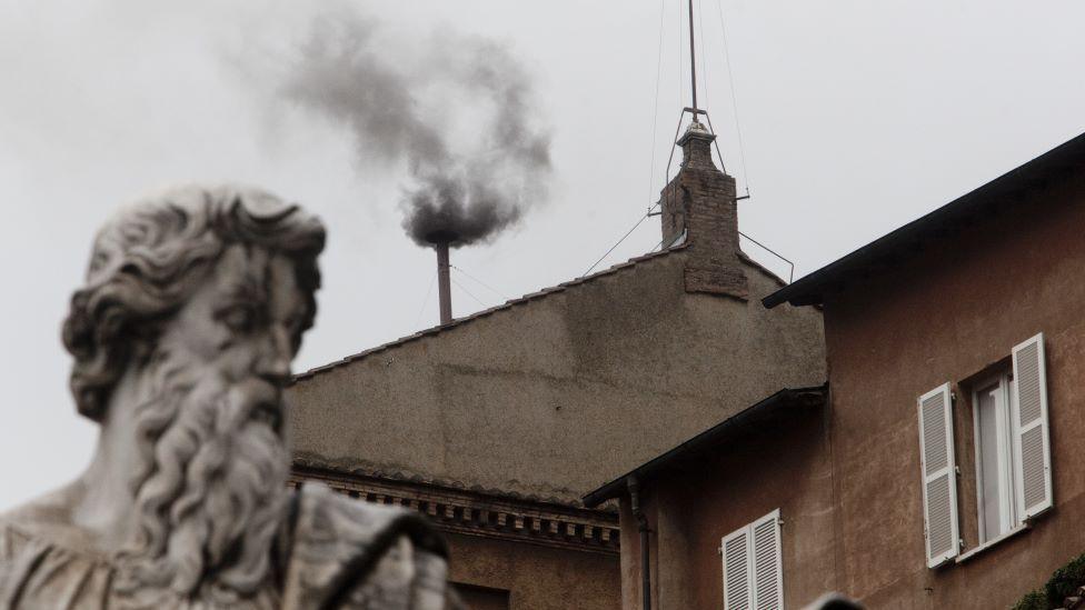 Black smoke billows out from a chimney on the roof of the Sistine Chapel, with a grey statue of a man in the foreground