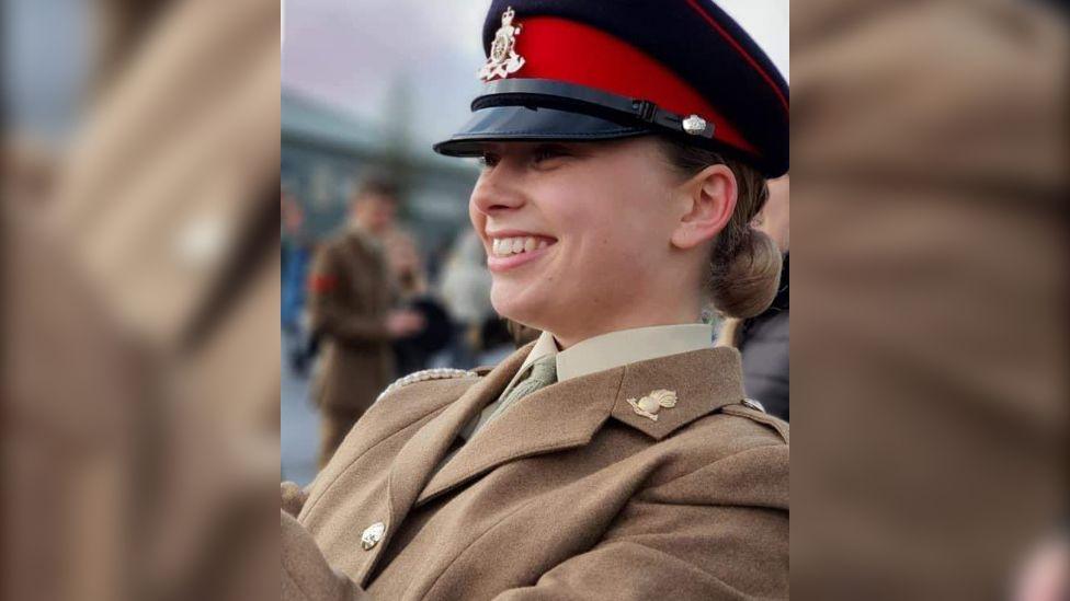 Jaysley Beck wearing her brown formal military uniform and a black and red cap. She has her light brown hair tied back into a low bun and is looking off to the left of the camera and smiling.