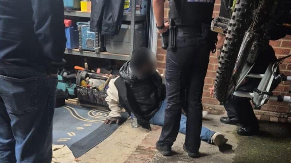 A garage with two people, one of them a police officer, standing above another person sitting on the floor, whose face is blurred,