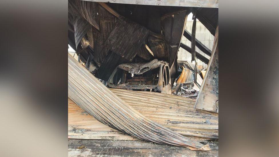 The remains of a building following a fire. Scraps of metal, which ones were walls, are collapsed on the ground. Inside you can see the remains of a burned-out car. 