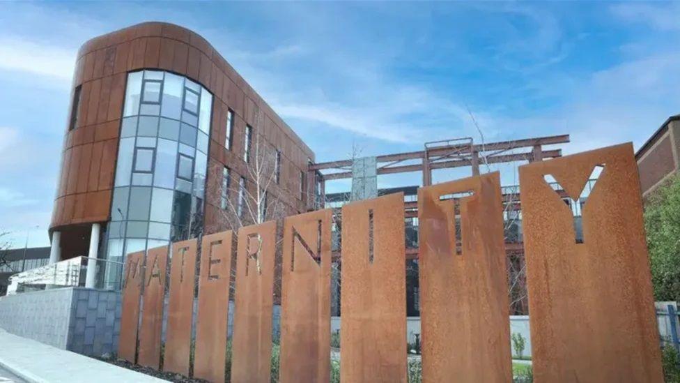 The new maternity hospital at the Royal Victoria site. There is a wooden fence with maternity on it in front of a modern wood and glass building.