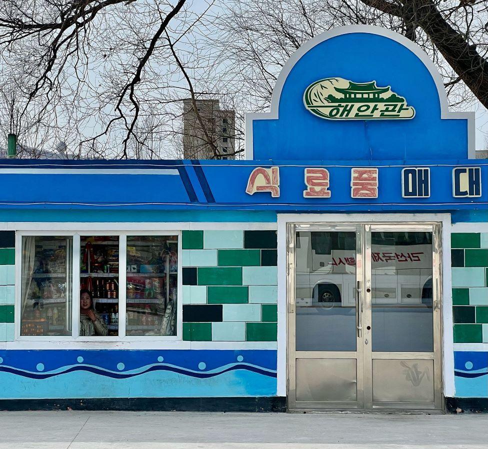 A photo of the exterior of a blue food store in North Korea. The exterior has green titles and there is a woman looking out of a window