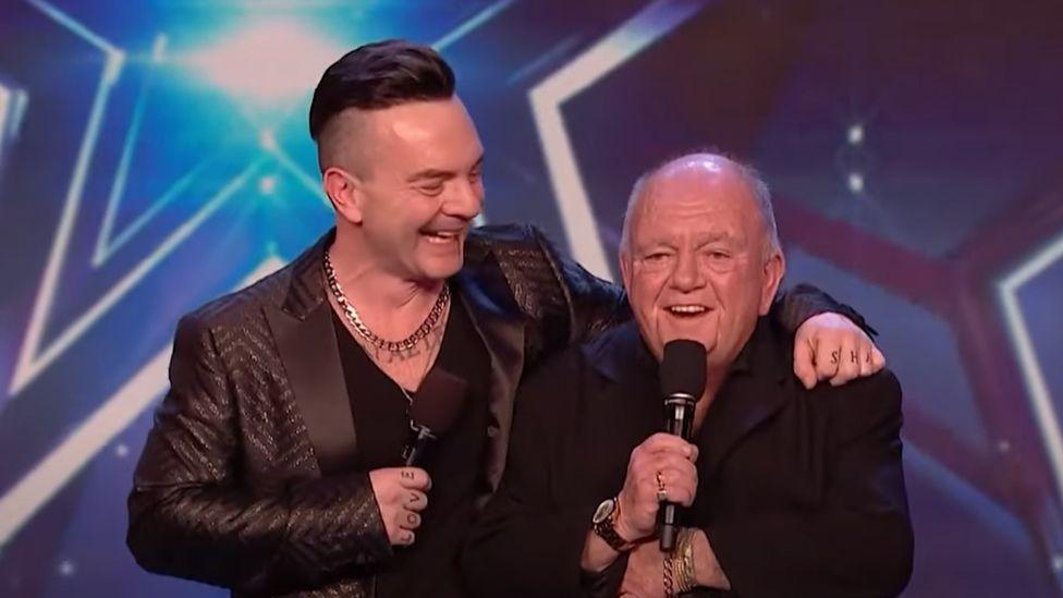 Dean, dressed as Robbie Williams in a black suit and t-shirt, stands on the Britain's Got Talent stage grinning with his arm round his dad's shoulder, who is also wearing a black jacket and holding a microphone. The background has a huge lit-up star.