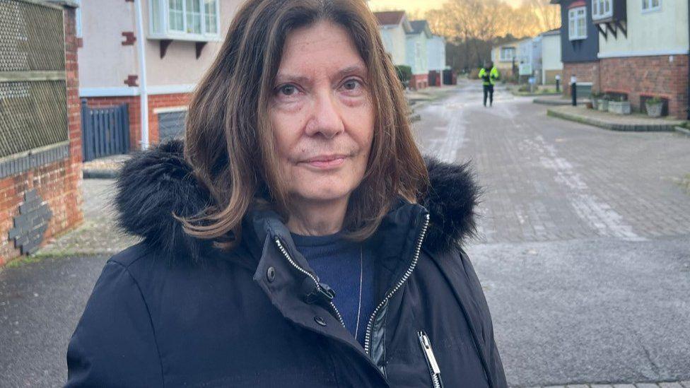 Woman with brown shoulder-length hair which is wearing a dark blue coat, rows of homes line both side of a road.