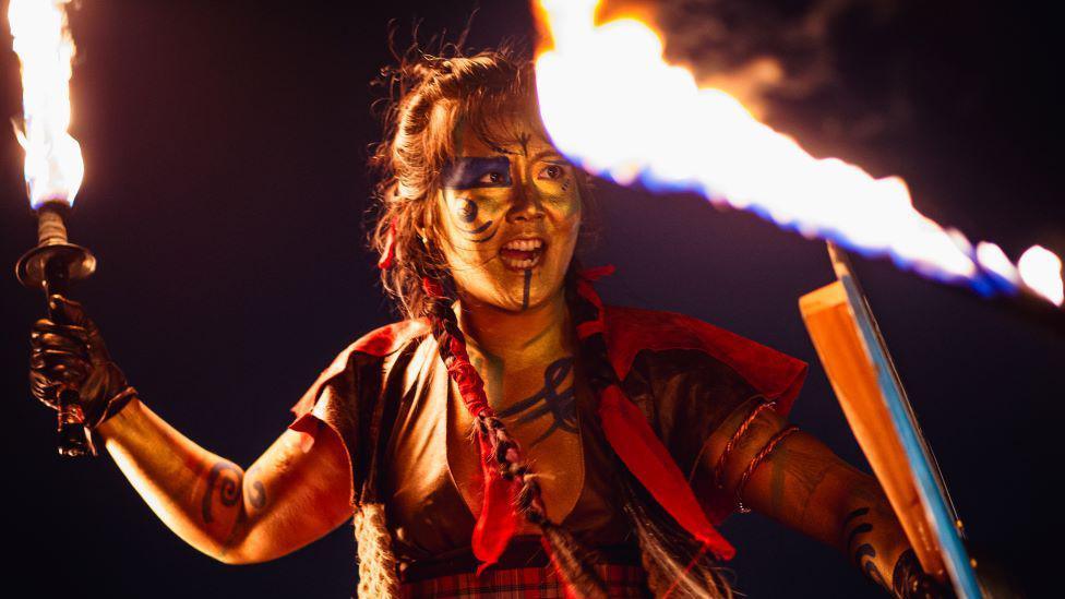 A woman with Celtic inspired face paint holding fire torch and shield.