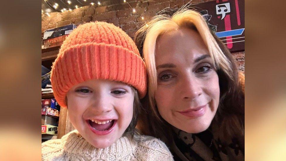 Milly on Air - aka Milly Chowles - posing for a selfie with her child, who is wearing an orange woolly hat