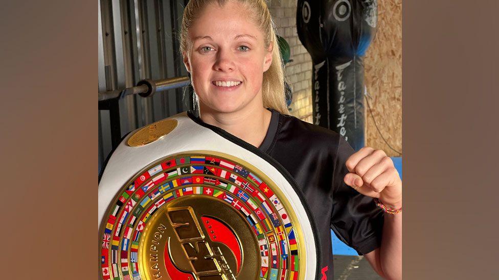 Hannah Turner, with fair hair pulled back into a high pony tail, wearing a black t-shirt, smiling, her left hand in a fist and the holding a colourful WKU K1 World Champion's belt