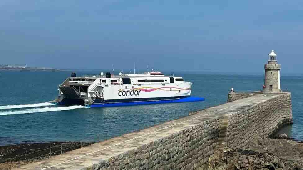 A ferry with the word Condor on the starboard flank motors put to sea with a stone harbour wall and a small lighthouse on the right and a cloudless sky above.