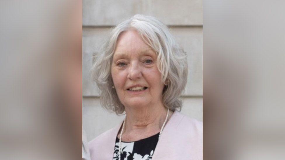 Pam, now in her 70s, with white shoulder-length hair. She is wearing a white and navy floral dress with a pink jacket and a string of pearls