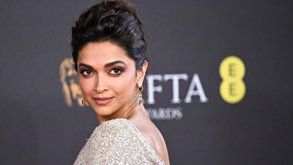 A woman with her hair in an updo wears dangly gold earrings and a backless, shimmering dress, looks over her shoulder at the viewer. Behind her a red carpet hoarding with the BAFTA logo can be seen.