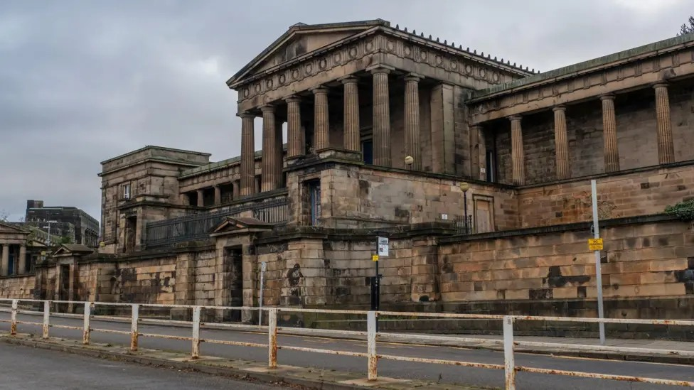 The neoclassical facade of the Old Royal High School, with a rusting white metal fence marking the road in the foreground and a leaden grey sky above.