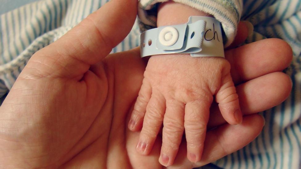 Newborn baby's hand, with a hospital identification wristband round it, being held by their mother
