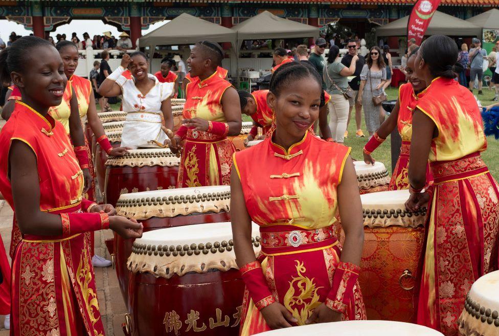 Women dressed in red, embroidered tops and skirts stand next to large ceremonial drums.