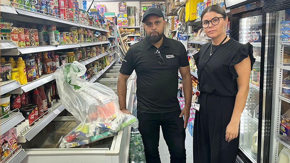 Nazir Ali and Samantha Veira stand in the aisle of a grocery store. Nazir is wearing a black polo shirt and dark pants, while Samantha is wearing a black dress. The aisle is filled with various food products on shelves, including canned goods, sauces, and snacks. A plastic bag filled with confiscated items sits atop a freezer in front of them.