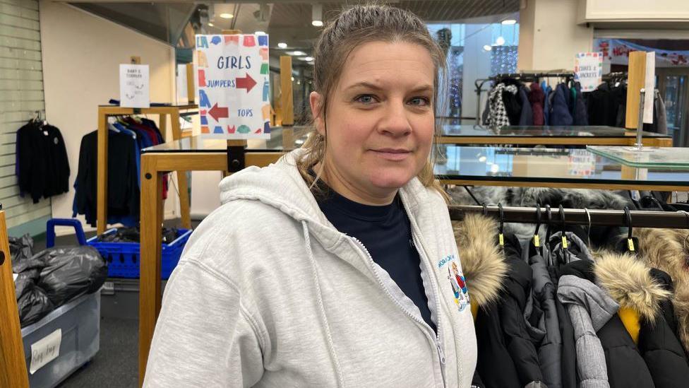 Moyra Younie - from Moray School Bank - smiling at camera, wearing a light grey hoodie and black top, with clothes on hangers in background.