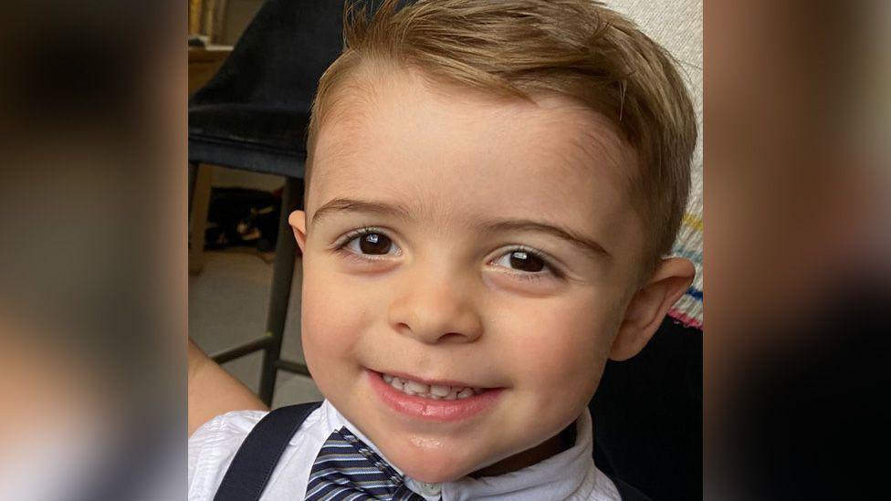 Three-year-old Freddie smiling at the camera, he has big brown eyes and sandy blonde hair