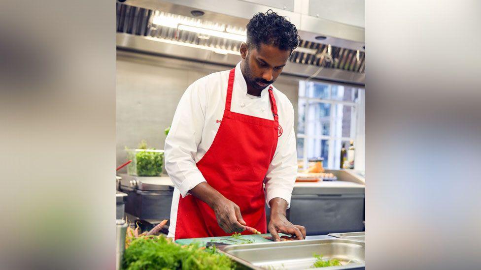 Brin Pirathapan he is cooking and wearing a red apron and white shirt