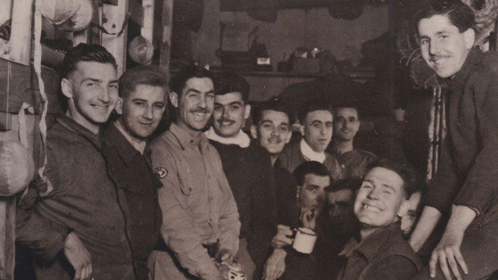 A black and white image showing a group of at least 12 World War Two Allied prisoners of war of the Japanese after liberation. They are all looking towards the camera and smiling broadly, standing in a hut, in a variety of uniforms. 