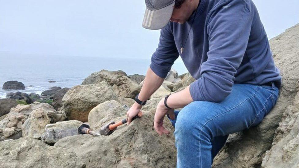 Man wearing baseball cap sat on a boulder with a hammer - the edge of a fossil can be seen on the face of a rock.