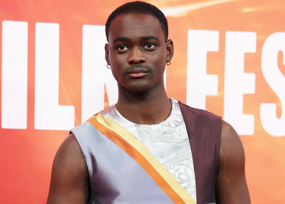 Ethan Herisse poses on the red carpet for the Nickel Boys gala screening at the BFI London Film Festival - he is wearing a silver, yellow, white and brown silk and lace sleeveless top.