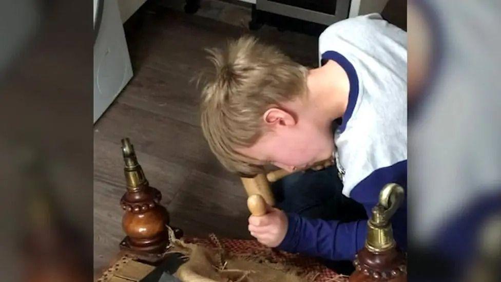 A blond boy looking closely at the under-side of of an ottoman while holding two tools