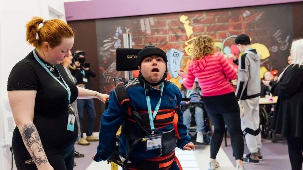 Young people take part in n activity at Corseford College, in a big room with graffiti artwork on the wall. A young man is in the foreground wearing a blue and orange jumper with a carer beside him.