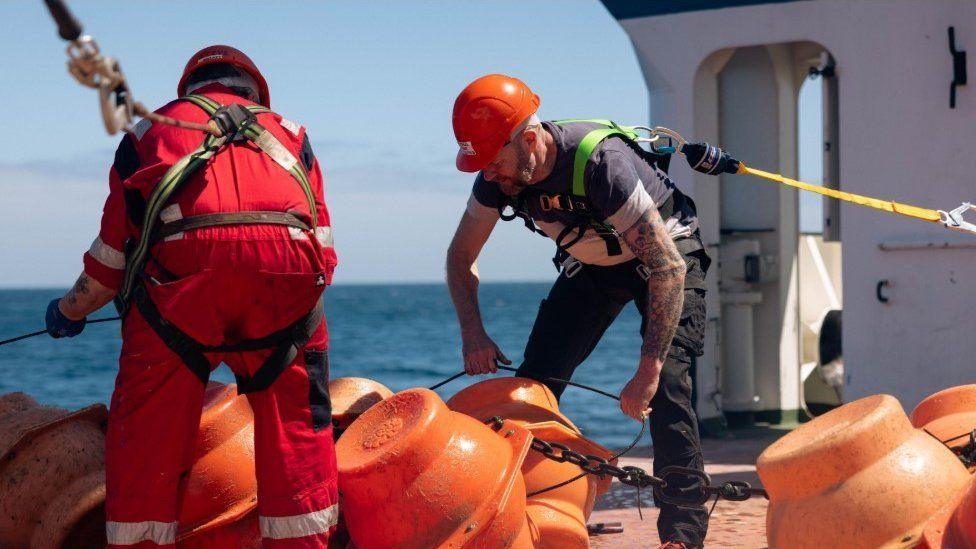 Two men in high vis and hard hats are sorting the moorings on the desk of RRS Discovery