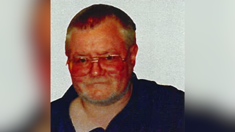 Mr Berryman is wearing glasses and a blue t-shirt. There's a white wall behind him.