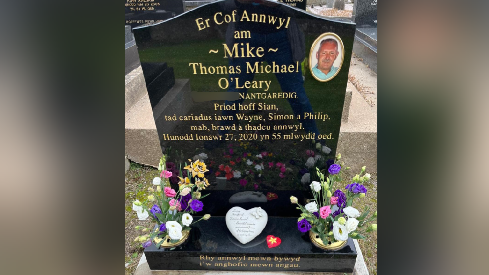 A black gravestone with a picture of Mike O'Leary, in a golden circle in the top righthand corner. The writing is in Welsh and reads: Er Cof Annwyl am Mike Thomas Michael O'Leary: Awel-Haf, Nantgaredig, Priod hoff Sian, tad cariadus Wayne, Simon a Philip, mab, brawd, a thadcu annwyl. Hunodd Ionawr, 27, 2020 yn 55 mlwydd oed