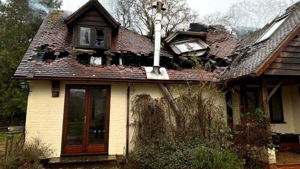 Brick house painted pale yellow with brown wooden doors and windows - there are gaping holes in the roof and smoke can been seen coming out.