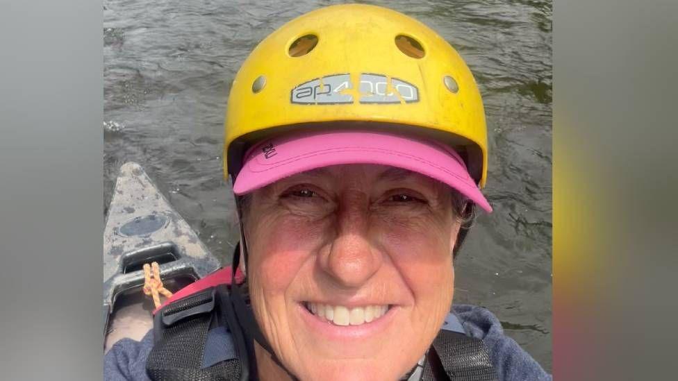 Wildlife filmmaker Justine Evans takes a selfie while canoeing on the river Wye.  She is sat in a black kayak, wearing a dark blue hoodie, a red life jacket and a pink baseball cap.  