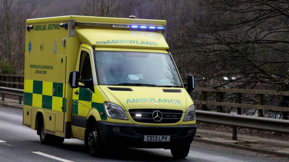 A picture of an ambulance driving down a road. The ambulance is yellow and can be seen with the Welsh word ambiwlans written on the front.