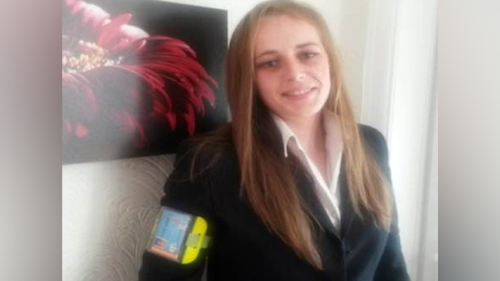 Chereiss Bailey is wearing a white shirt and black jacket and has long, light brown hair. She is leaning towards a wall and smiling at the camera. There is a picture on the wall behind her.
