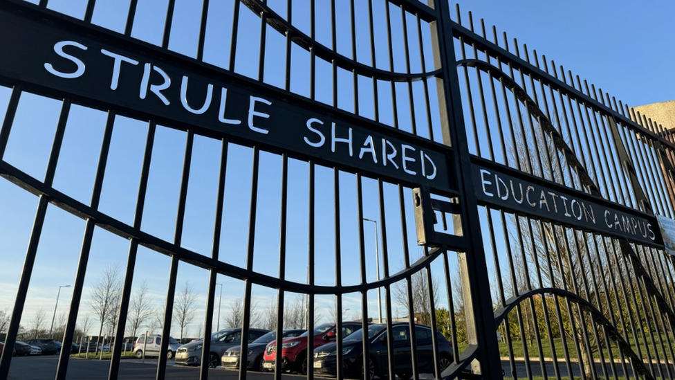 A number of parked cars are seen in a car park through gates which have a Strule shared education campus sign across them