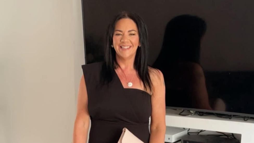 A woman standing in front of a TV and white wall, she is wearing a black dress and has black hair