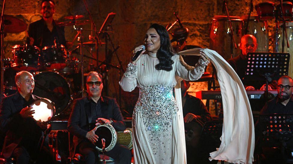 Ahlam performs on stage while wearing a white dress with jewels on it. Behind her is a band, including men in black suits playing a drum, tambourine and drums - Thursday 21 August 2025.