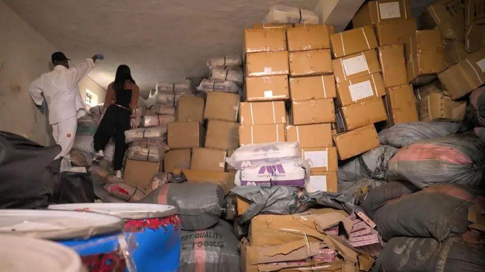 Piles of cardboard boxes and large packages wrapped in plastic stacked up to the ceiling inside a warehouse.