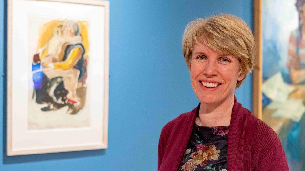 A woman standing in a museum. She has thick short blonde hair, swept over her forehead, and is smiling. To her right is an out-of-focus Rosemary Rutherford painting, of a child hugging an adult. The walls of the gallery are bright blue.