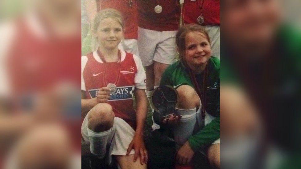 A photo of a young Rhiannon aged about 12, smiling to camera, wearing football kit and kneeling down amidst other players, holding a trophy and wearing a medal