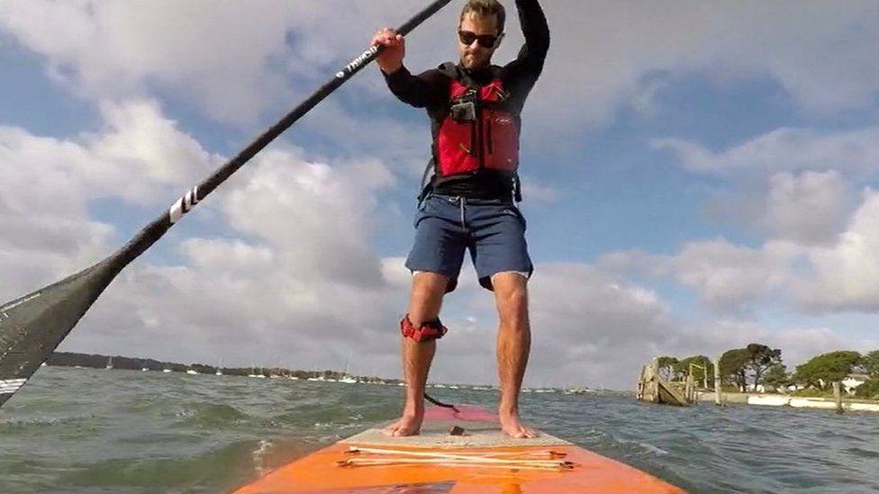 Orange paddleboard on water with man in shorts standing on it he is wearing a life jacket and has a black paddle in the water on the left side.