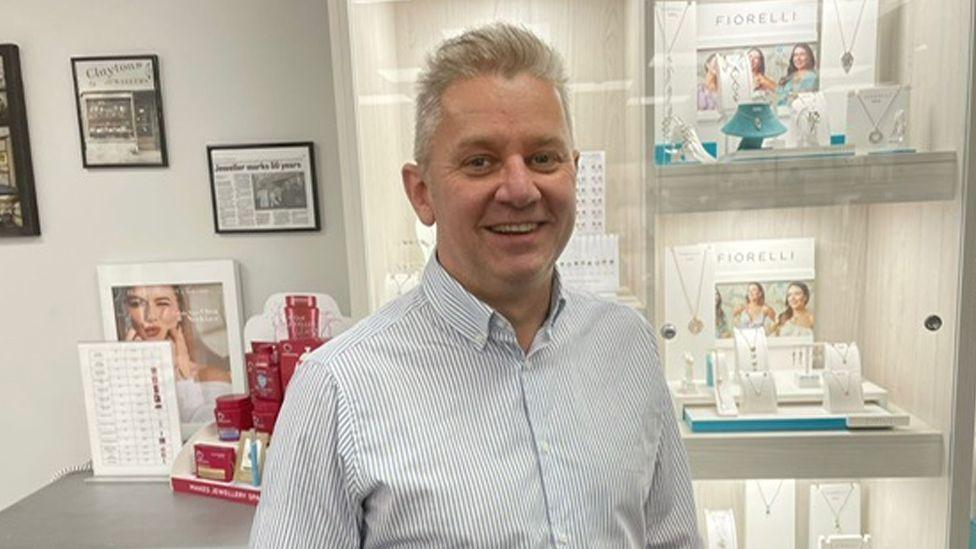 Jeweller Andrew Clayton standing inside his shop in front of jewellery display cases. He is smiling and has short grey hair and is wearing a striped white and blue shirt