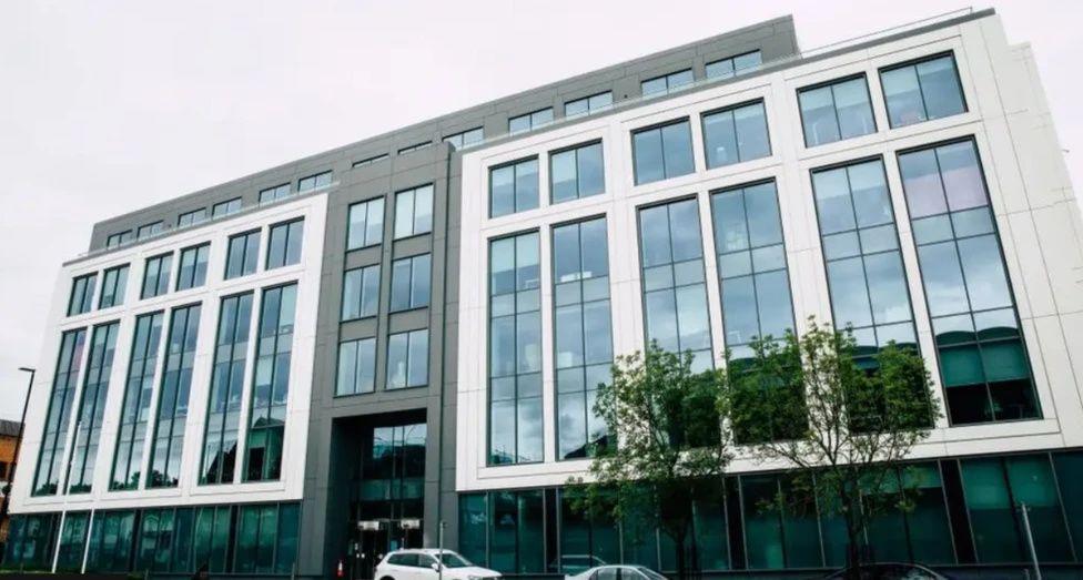 Grey and white office building with large glass windows and two green trees outside.