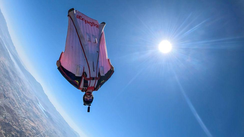 Man - Liam Byrne - wearing wingsuit, upside down in mid air and smiling at camera, with sun in sky.