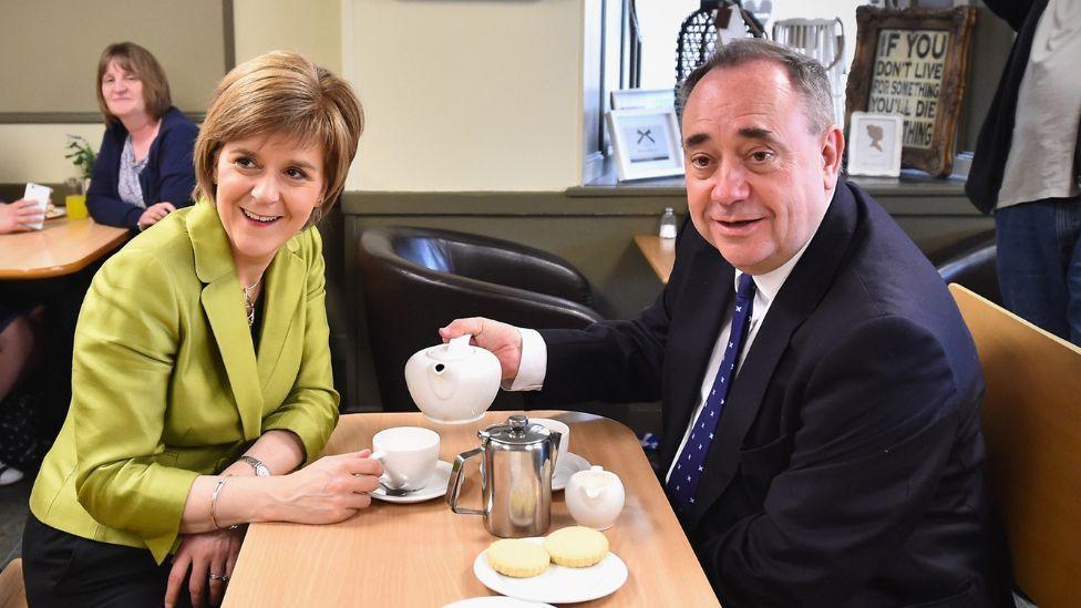A man with thinning dark hair and a woman with short fair hair sit opposite each other at a table in a cafe. The man, on the right, is holding a pot of tea above the woman's cup. He is wearing a dark suit, she is wearing a light green jacket. They are both smiling. There are biscuits, a coffee pot and a milk pourer on the table. 