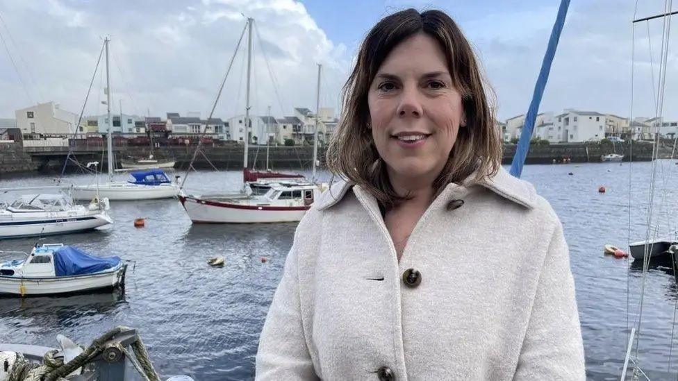 A woman with short brown hair, wearing a white coat, is smiling at the camera. She is standing in front of Porthmadog harbour. 