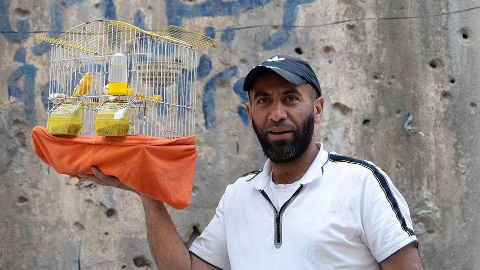 Yasir stands outside a bullet-marked building holding a cage containing his pet bird