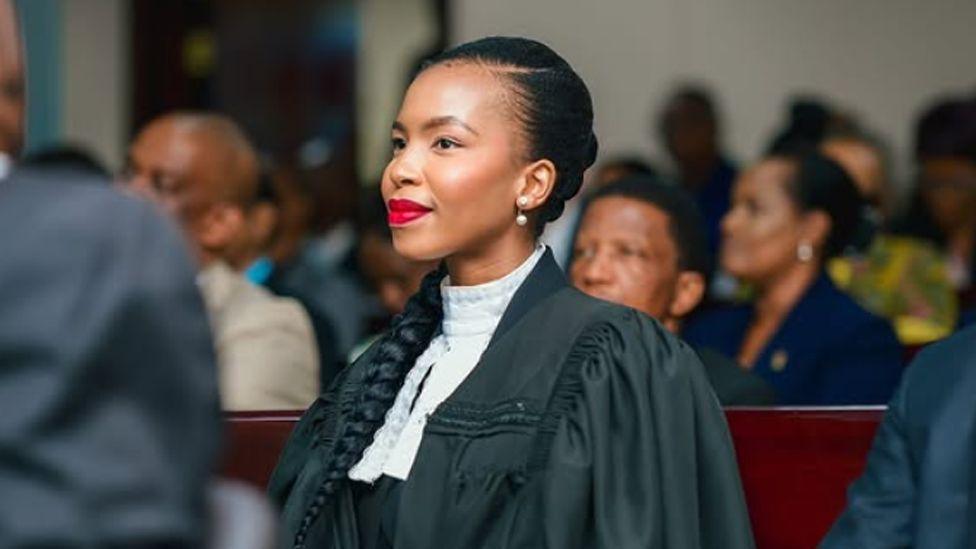 Lesego Chombo in a black legal gown and white collar. Wearing red lipstick, she is seated on a bench with others in a courtroom and is looking to the side - not at the camera.